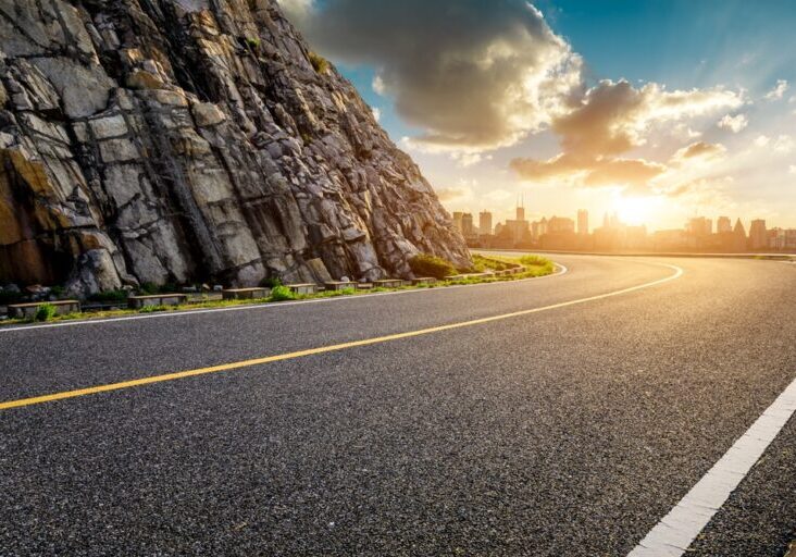 Asphalt,Highway,Road,And,Mountain,With,City,Skyline,At,Sunset